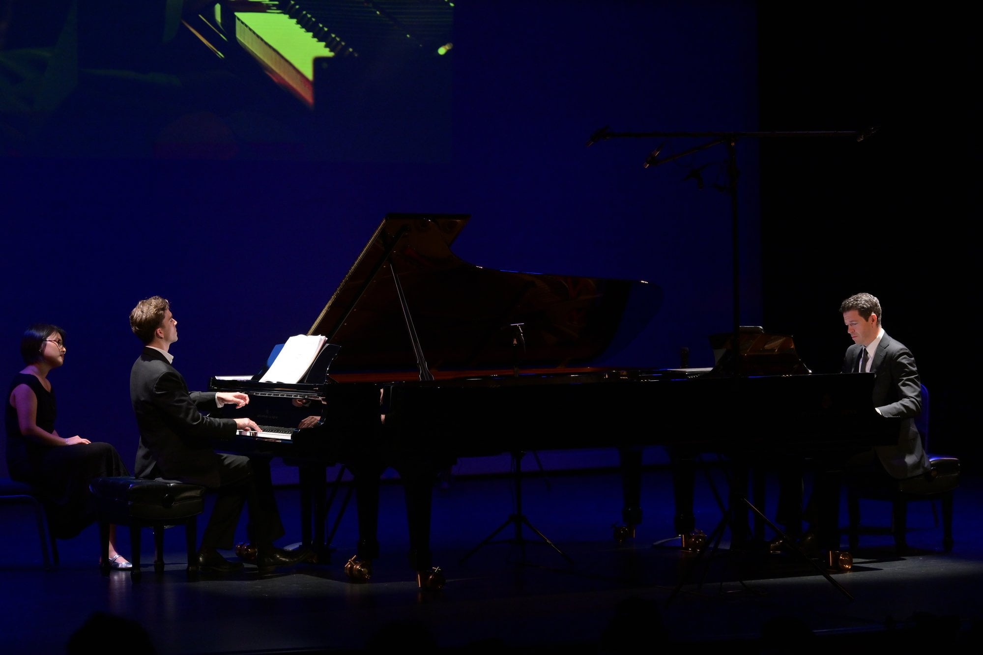 Two pianos on a blue-lit stage, with a page turner beside the first piano